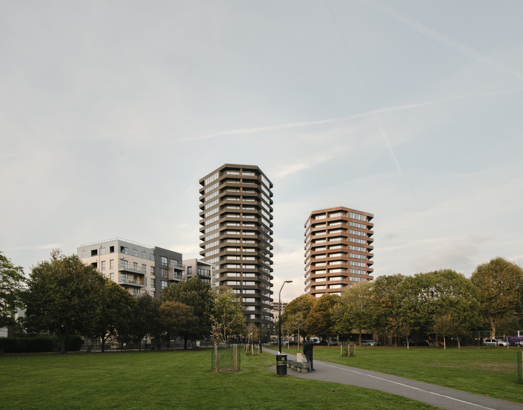 Two medium rise residential towers, one red and the other grey, overlook a tree-filled park.